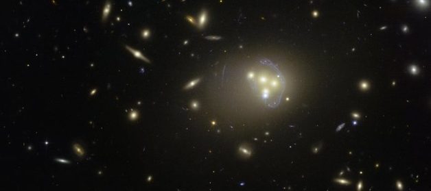 Image - This image from the NASA/ESA Hubble Space Telescope shows the galaxy cluster Abell 3827. The blue structures surrounding the central galaxies are views of a more distant galaxy behind the cluster that has been distorted by an effect known as gravitational lensing. Observations of the central four merging galaxies in this image have provided hints that the dark matter around one of the galaxies is not moving with the galaxy itself, possibly indicating the occurrence of an unknown type of dark matter interactions. (Credit: ESO)