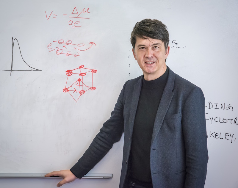 Gerd Ceder, a person with short dark hair wearing a black top and charcoal blazer, leans against a whiteboard.