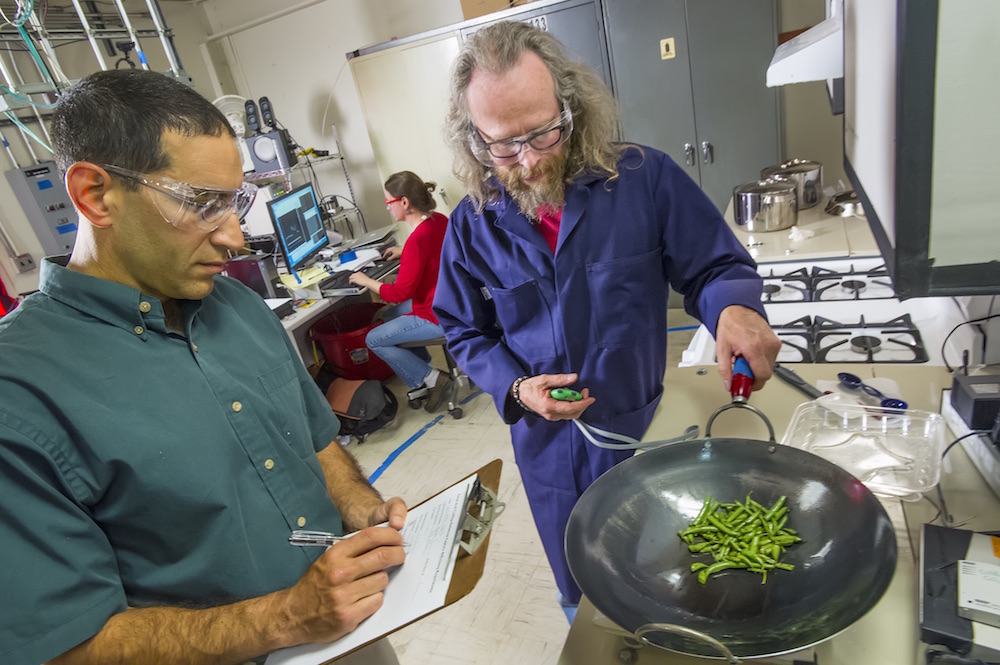 One researcher fries green beans on a fry pan indoors as another takes notes.
