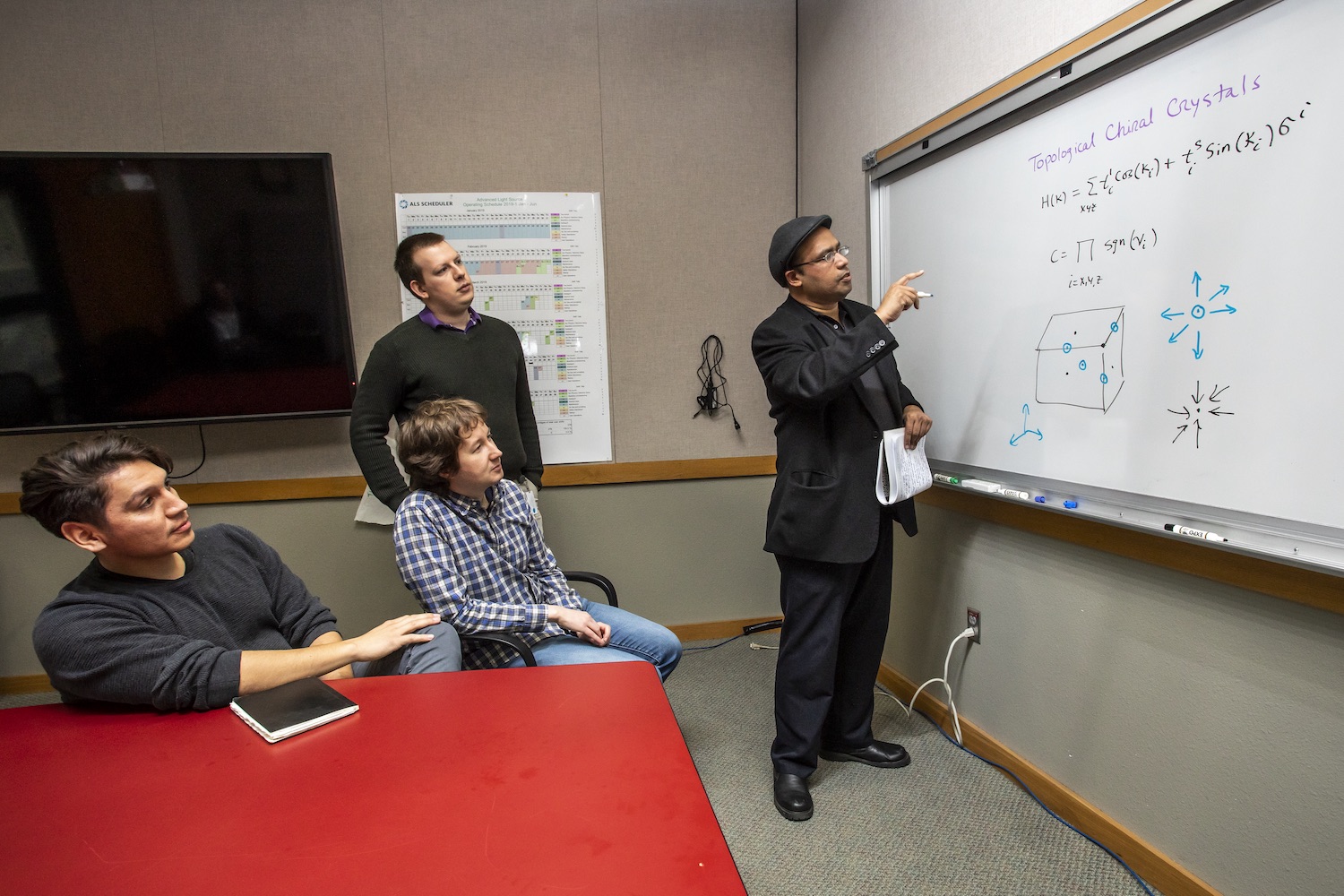 Image - Princeton University Professor Zahid Hasan, right, describes the exotic behavior of electrons in topological crystals that were studied at Berkeley Lab. Members of Hasan’s research team observe, including: Daniel S. Sanchez (left), Ilya Belopolski (standing), and Tyler A. Cochran, (seated, second from left). (Credit: Marilyn Chung/Berkeley Lab)