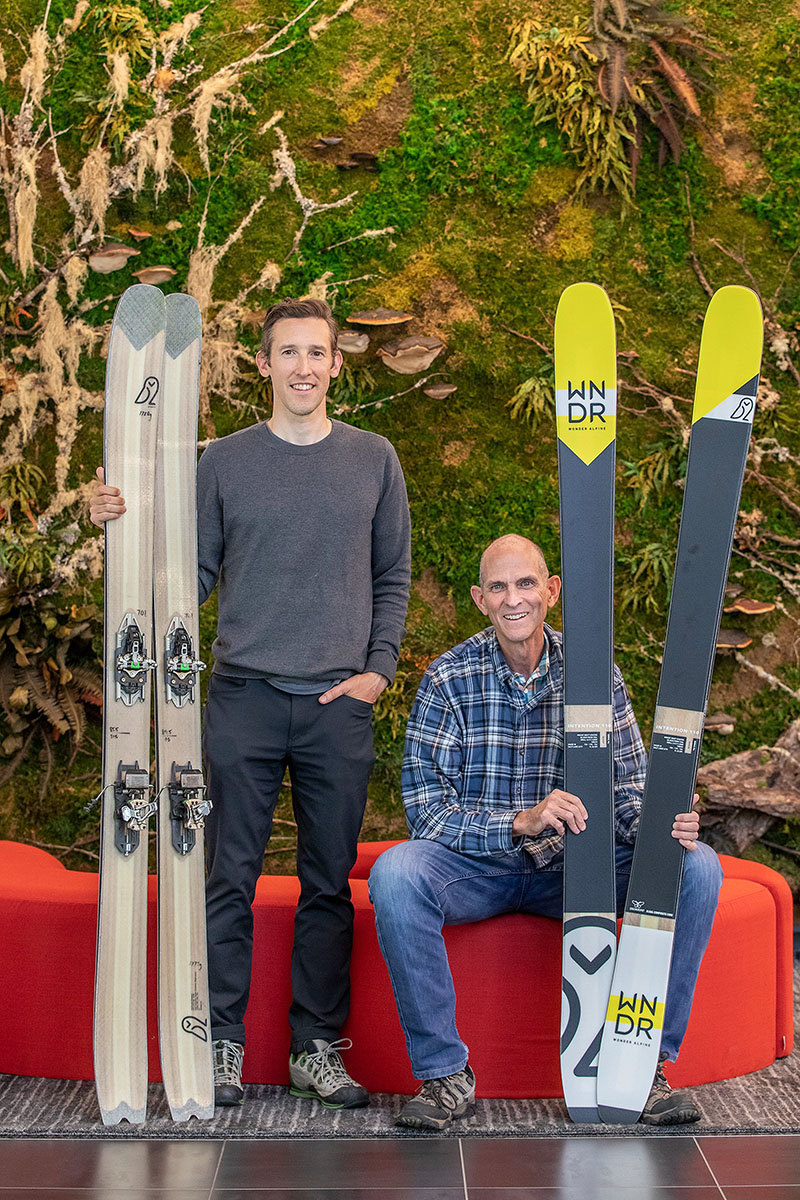 Two people in front of a red couch holding skis. 