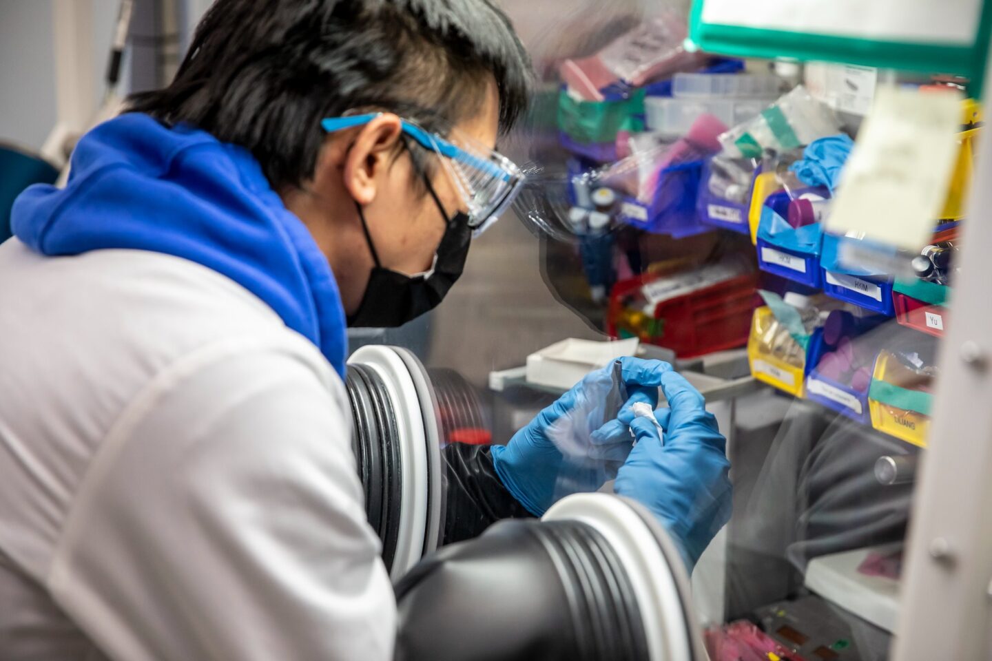 A scientist in a white coat and blue hoodie holds a DRX sample in a fume hood.