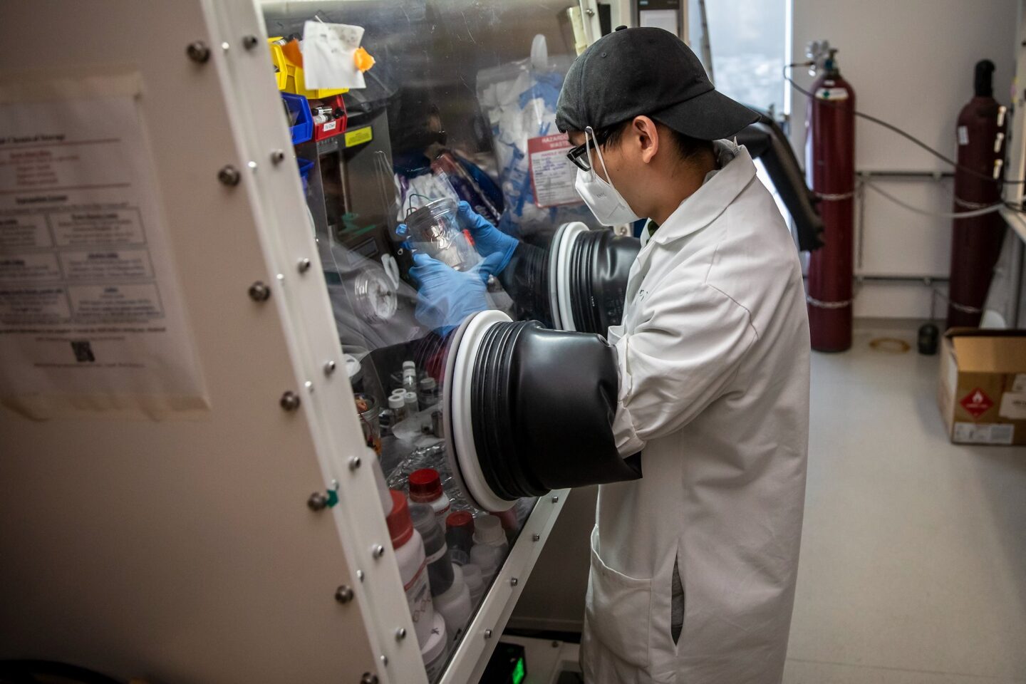 Scientist in a white coat and black backwards baseball cap handles a sample in a fume hood.