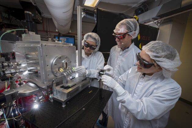 Three scientists in white lab suites inspect an accelerator.