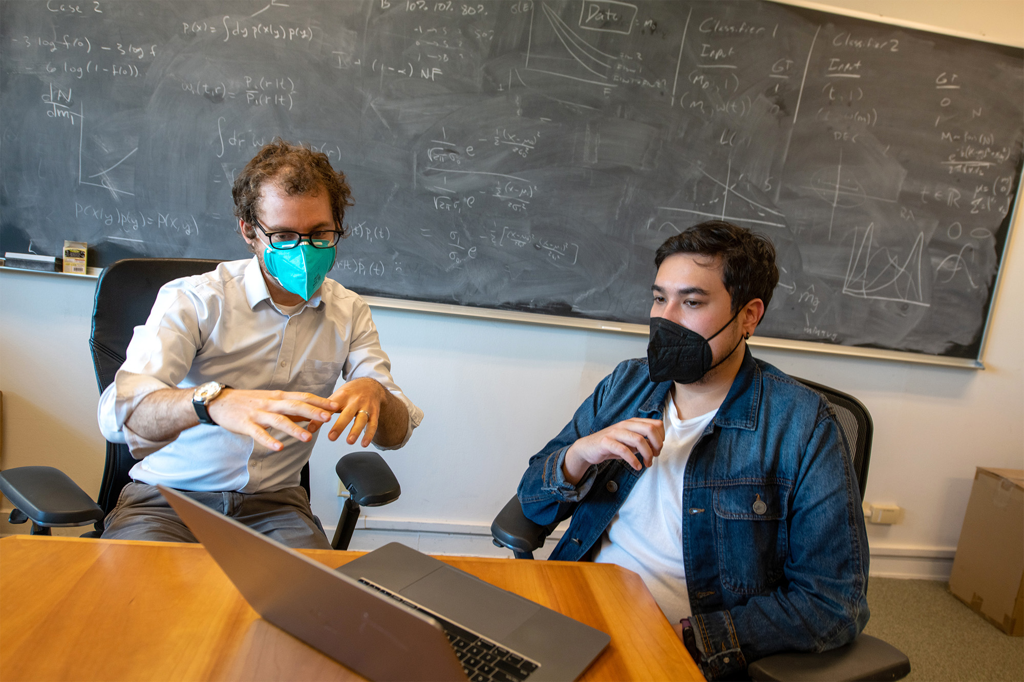 Two people wearing face masks in discussion while looking at a laptop. In the background is a chalkboard with calculations.