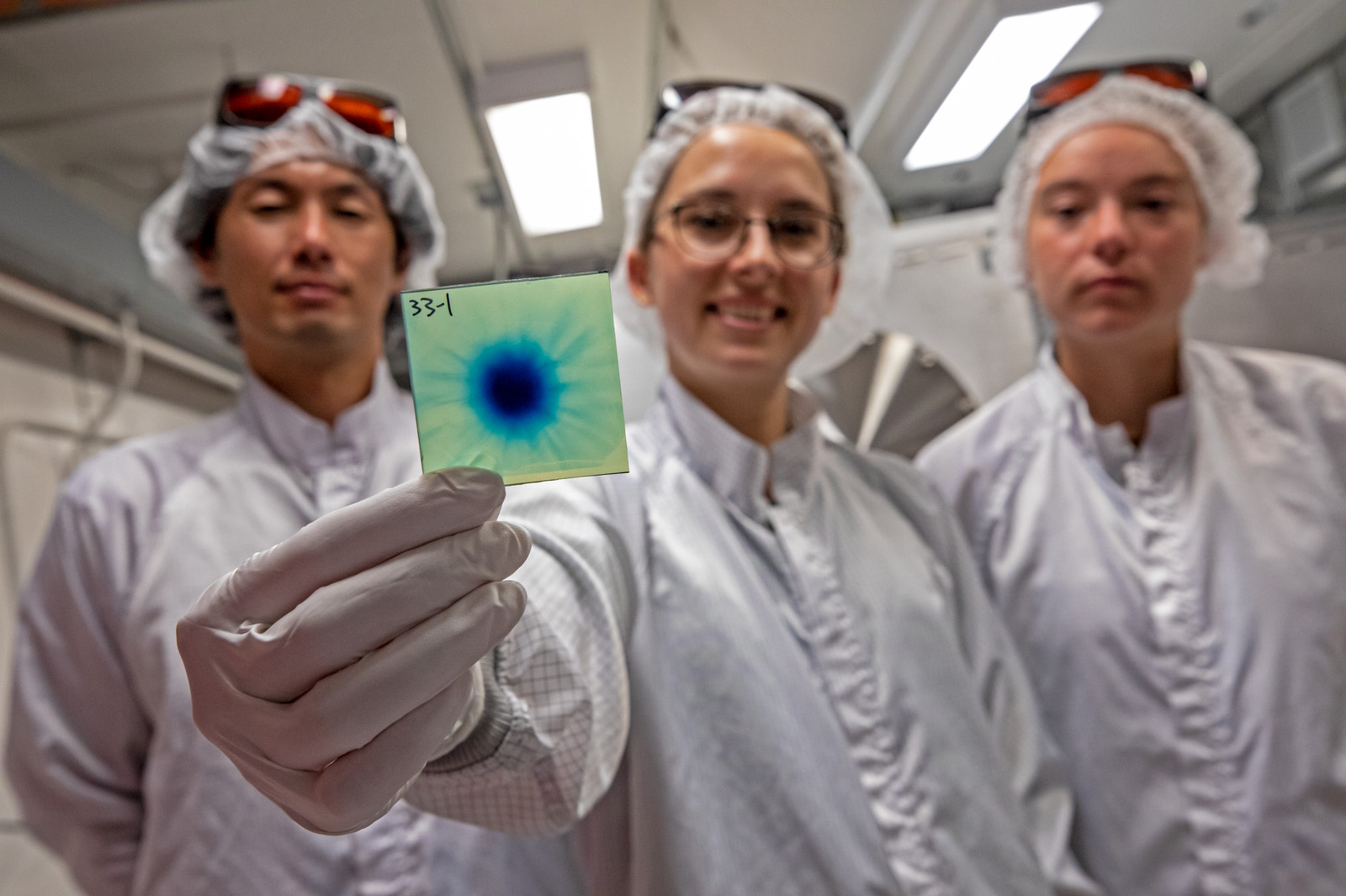 Three scientists in protective gear hold up a green and blue radiochromic film.