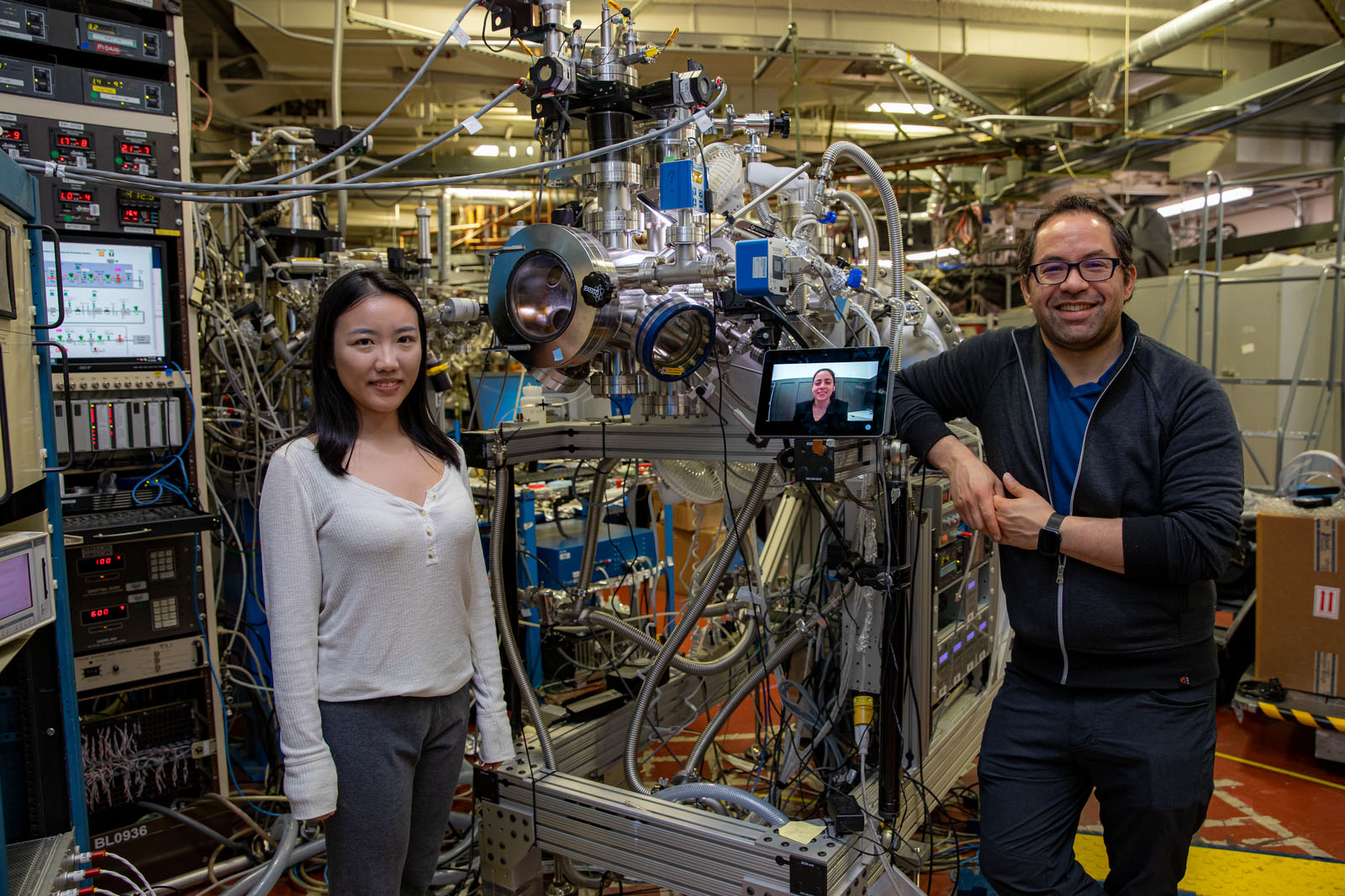 Two scientists pose with another scientist on Zoom next to their scientific equipment in the Advanced Light Source.