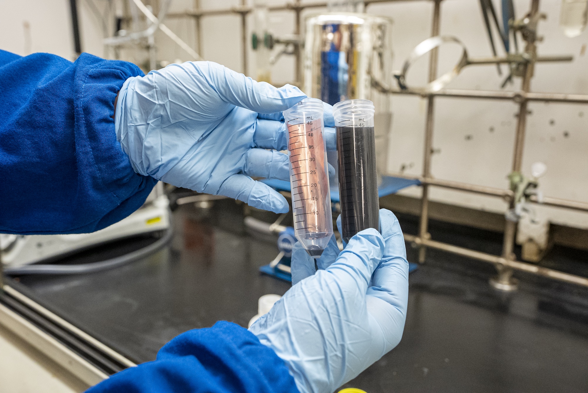 Two hands with blue gloves are golding tubes in a lab setting. One tube is filled with a piece of copper that has been cleaned of other components and the other with the alkaline solution used to dissolve the quick-release battery binder