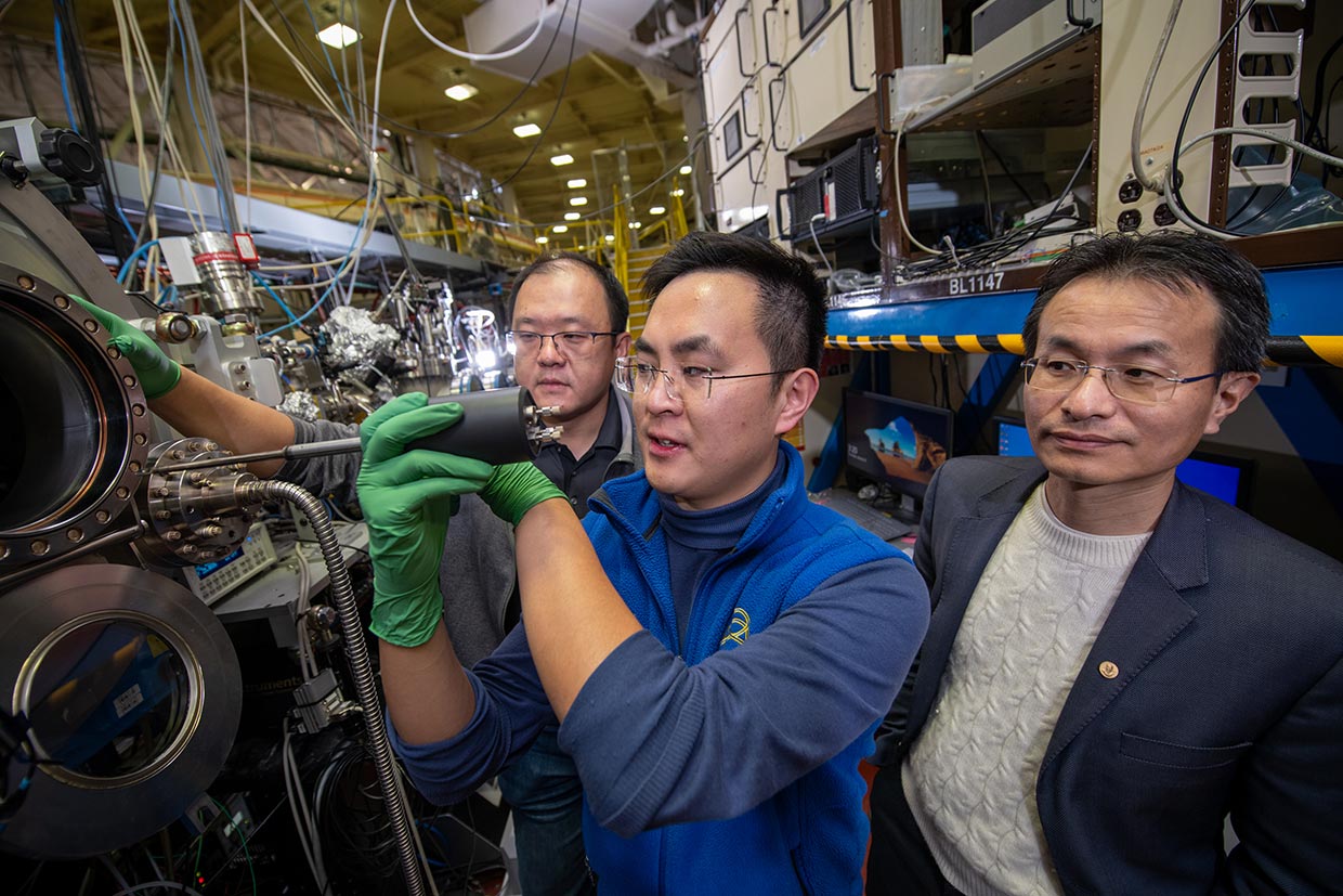 One scientist loads a sample into the soft X-ray scattering chamber as two other scientists to the left and right observe at the RSoXS Beamline at the Advanced Light Source.