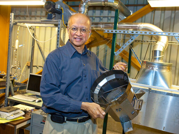 Ashok Gadgil, a person with short gray hair wearing glasses and a navy collared shirt, holds up a Berkeley-Darfur Stove in the lab.