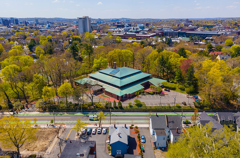Birdeye view of a city-scape. A large green roof dominates the center of the frame. Trees and forested area surround the building.