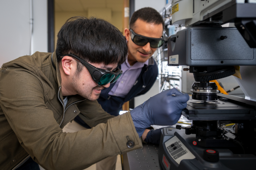 Two scientists with short black hair prepare some samples on a spectroscopy instrument that is on the right of the frame.