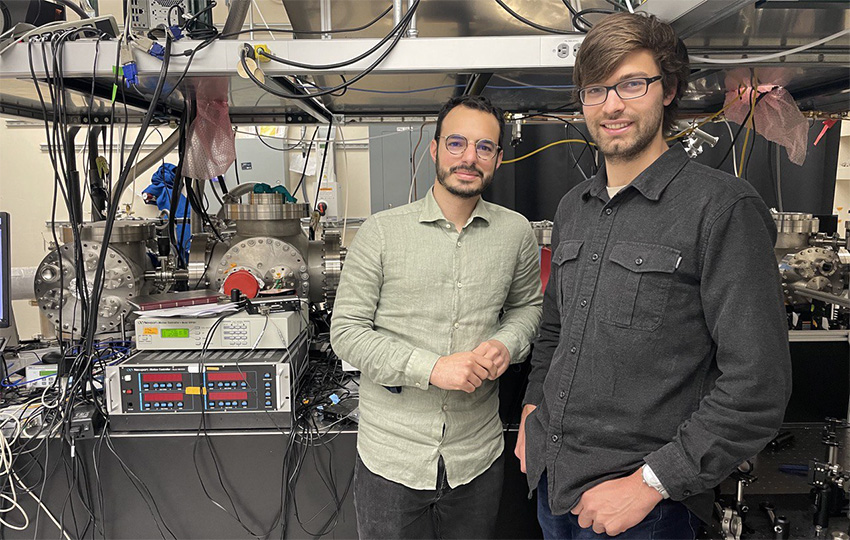 Enrico Ridente, a person with short dark hair and a beard wearing a light green collared shirt and black pants, stands next to Eric Haugen, a person with short brown hair wearing a dark collared shirt with jeans. The two are standing in front of a machine in their lab.