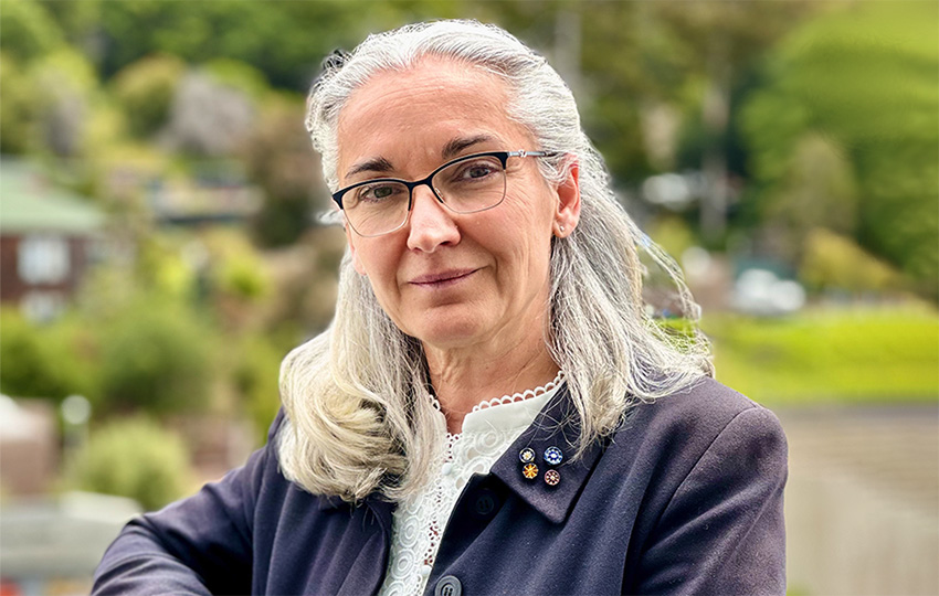 Eva Nogales, a person with medium-length white hair wearing glasses and a dark jacket over a white collared top, photographed outdoors with trees in the background.