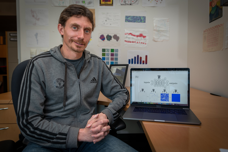 A staff scientist is photographed at his desk. Next to him is a laptop displaying computer simulations.