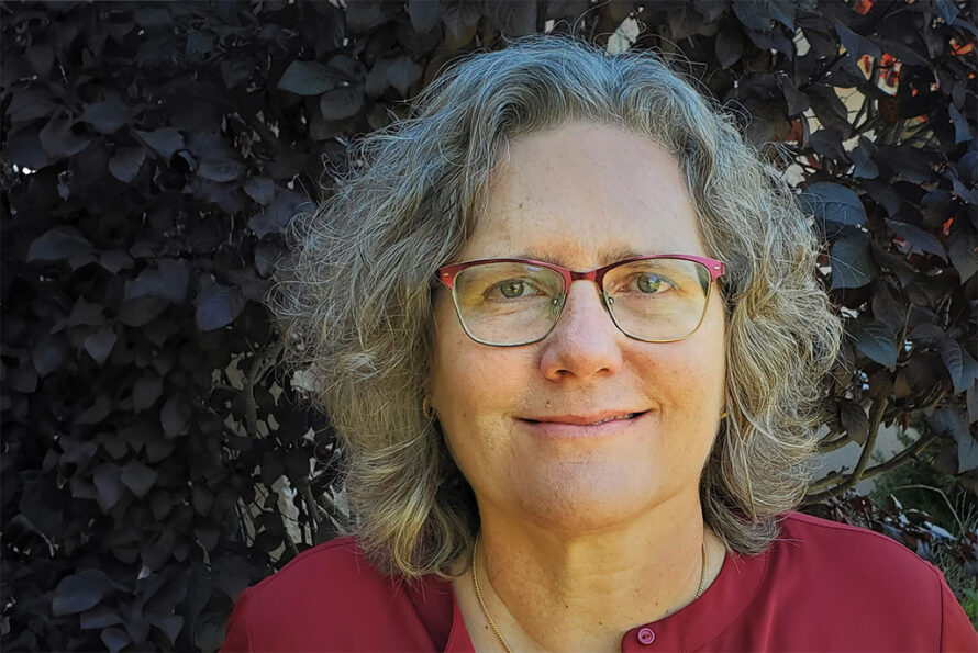 Lori Zscherpel, a person with shoulder-length gray hair wearing red framed glasses and a red top against a dark leafy background.