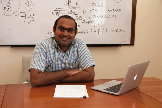 Kranthi Mandadapu, a person with short dark hair wearing glasses and a white and blue checkered shirt with arms folded on a desk with a laptop and papers. Behind him is a whiteboard with figures and calculations.