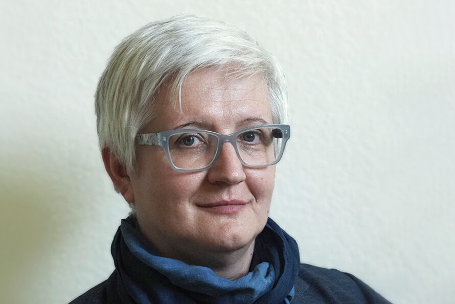 Ana Kupresanin, a person with short white hair with glasses wearing a blue top, photographed in front of a cream-colored wall.