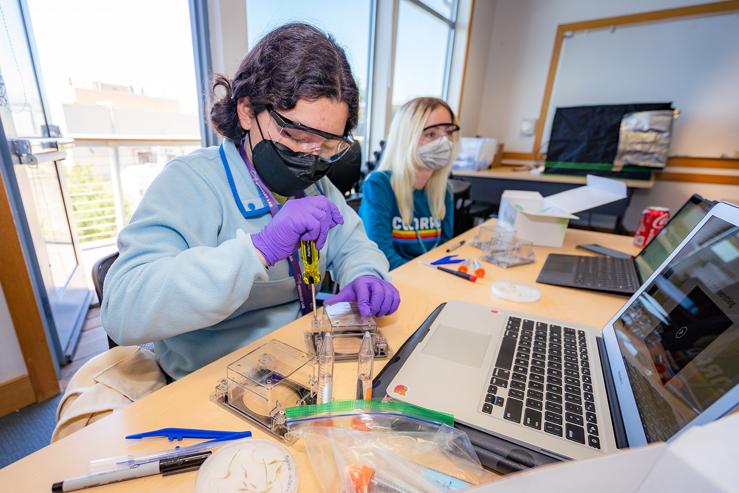 A masked student with dark hair, glasses, and purple latex gloves uses a screwdriver to put together an EcoFAB device.