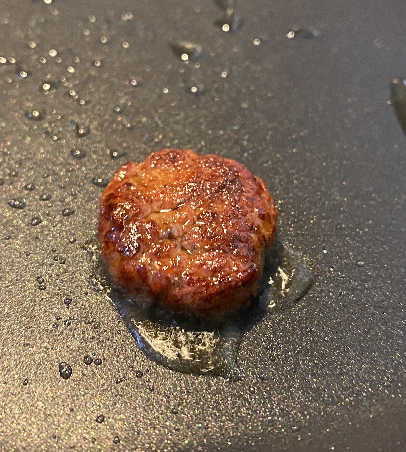 A small, rounded reddish mass that looks like a meat patty sitting in a frying pan beaded with oil