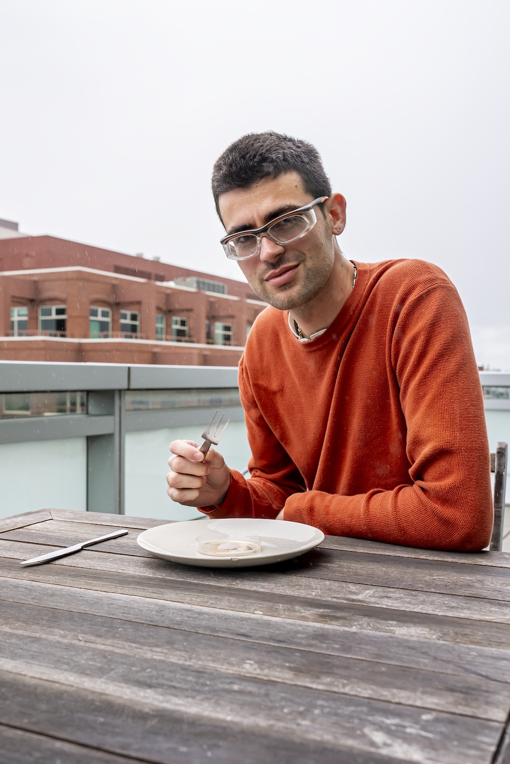 A young man in a long sleeve red top sitting outside on a picnic bench. He is wearing lab goggles and holding a fork. On the table in front of him is a plate containing a petri dish filled with fuzzy mold.