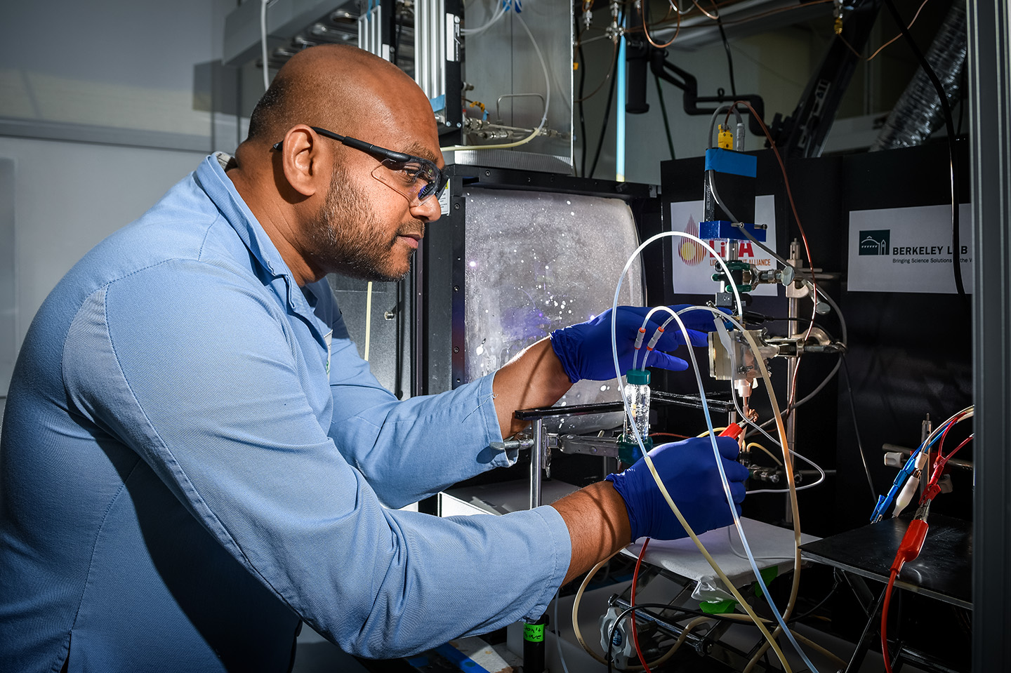 A researcher wearing safety goggles and blue gloves works with intricate lab equipment, adjusting tubes and wires in a lab setting.