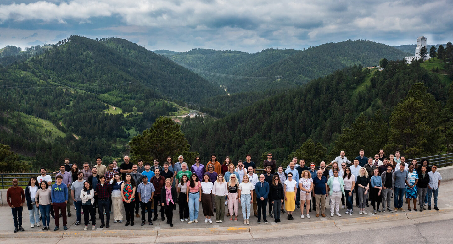 Group photo of the LZ collaboration members at the Sanford Underground Research Facility, taken outdoors with green mountains in the background.