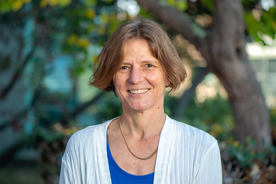 Daniela Leitner, a person with short brown hair wearing a white cardigan over a blue top and a gold necklace, photographed outdoors.