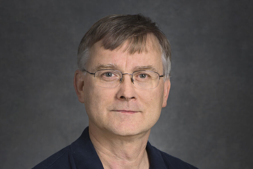 Portrait of Peter Fischer, a person with short brown gray hair wearing glasses and a dark top, photographed against a gray backdrop.