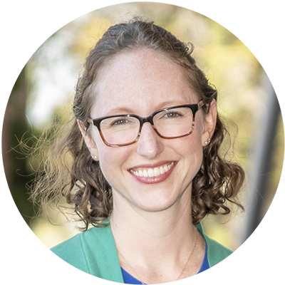 Portrait of Corinne Scown, a person with shoulder-length curly brown hair wearing glasses and a green top, smiling.