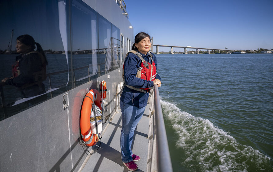 A scientist wearing a orange life vest leaning against the rails of a large research boat.