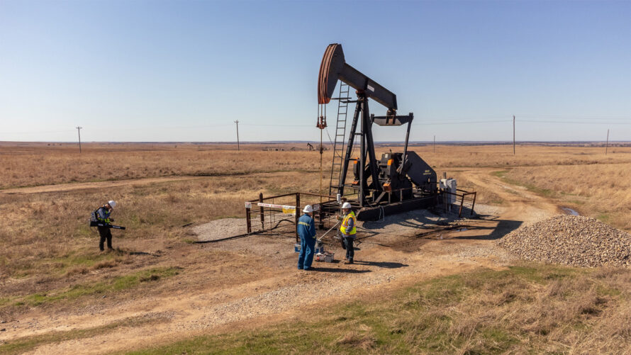 Oil pump jack in a field with people in hard hats around it.