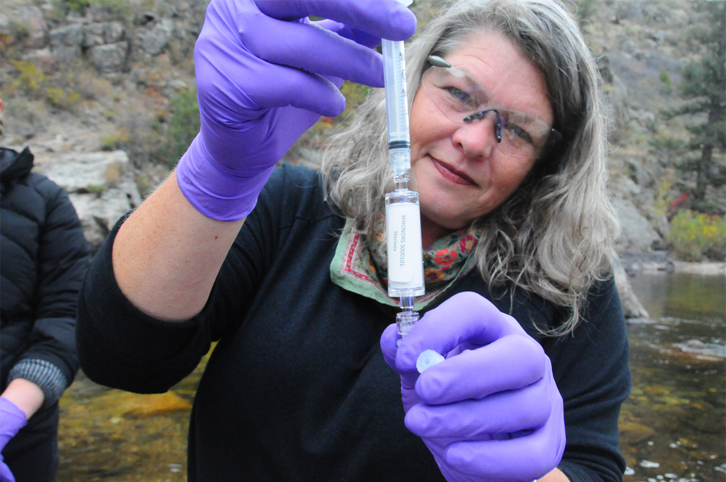 A person with medium-length gray hair wearing glasses and purple gloves, taking a sample outdoors.