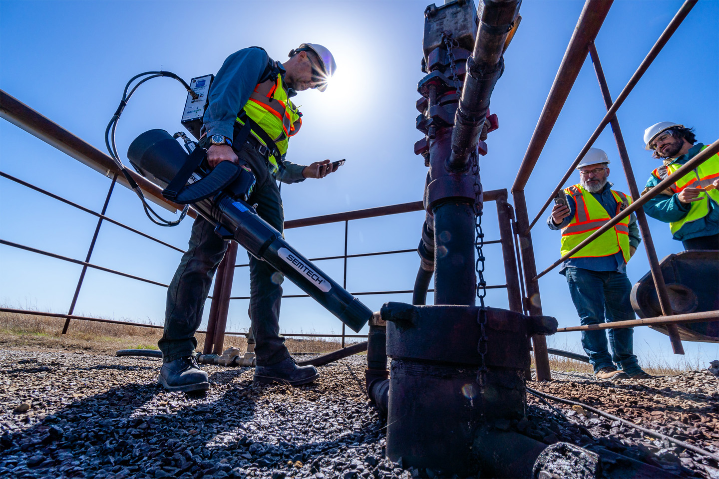 Three people in a hard hat and safety gear holding an emission quantification tool outdoors.