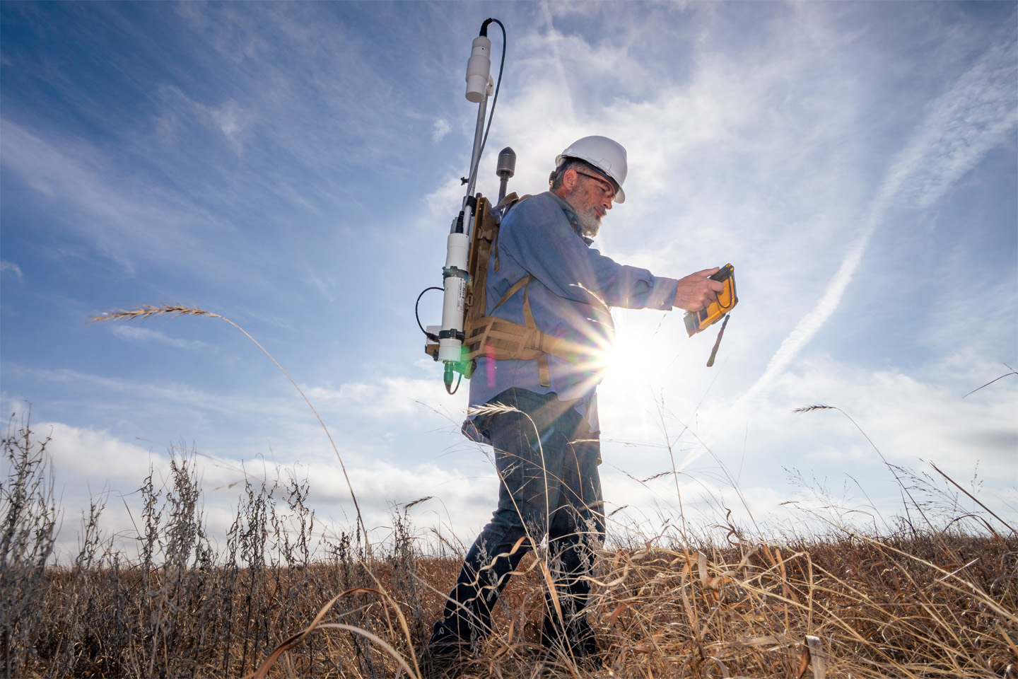 Scientist wearing a hard hat and backpack with a sensor on it, outdoors.