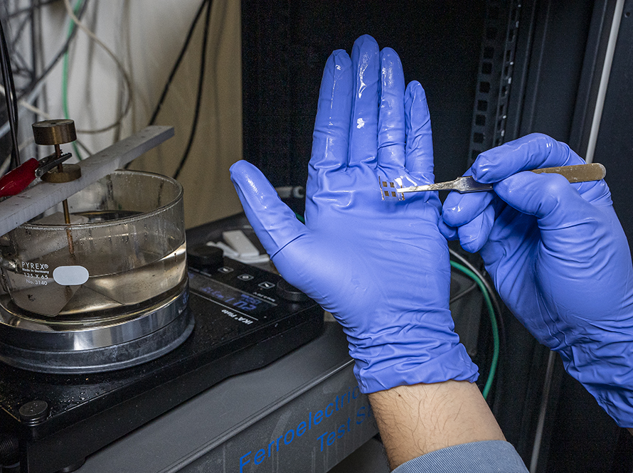 A gloved hand holding a small electronic component near a beaker of liquid in a lab setting.