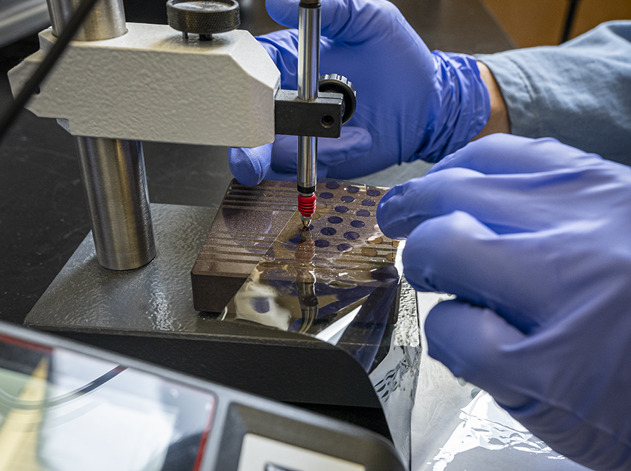 A gloved hand operates a precision tool, working with a sample on a flat surface in a laboratory environment.