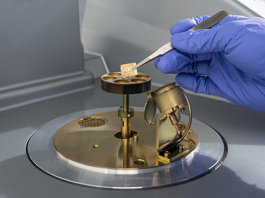 A gloved hand uses tweezers to place a small sample into a scientific instrument with metallic components.