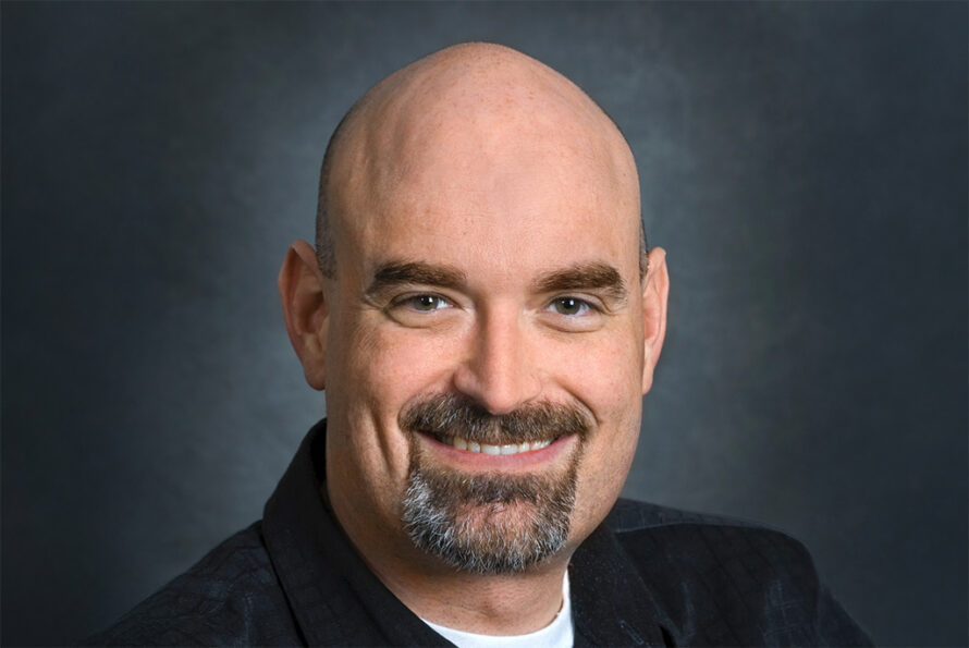 Portrait of a person with a gray beard smiling, wearing a dark jacket over a white top, photographed against a gray backdrop.