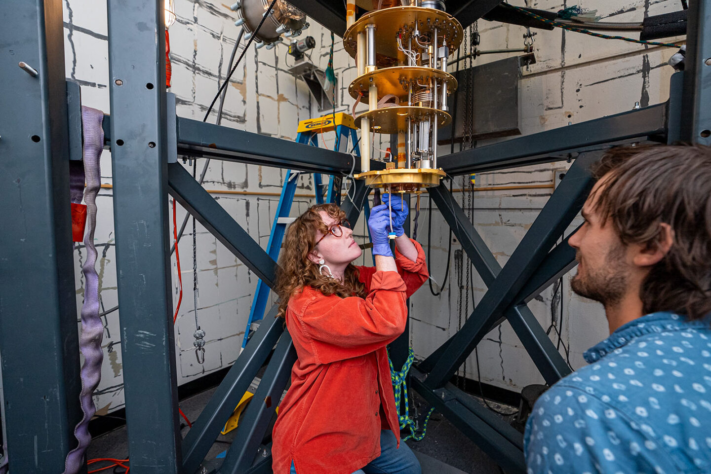 One researcher wearing purple gloves works on TESSERACT's dilution refrigerator, as another researcher watches.