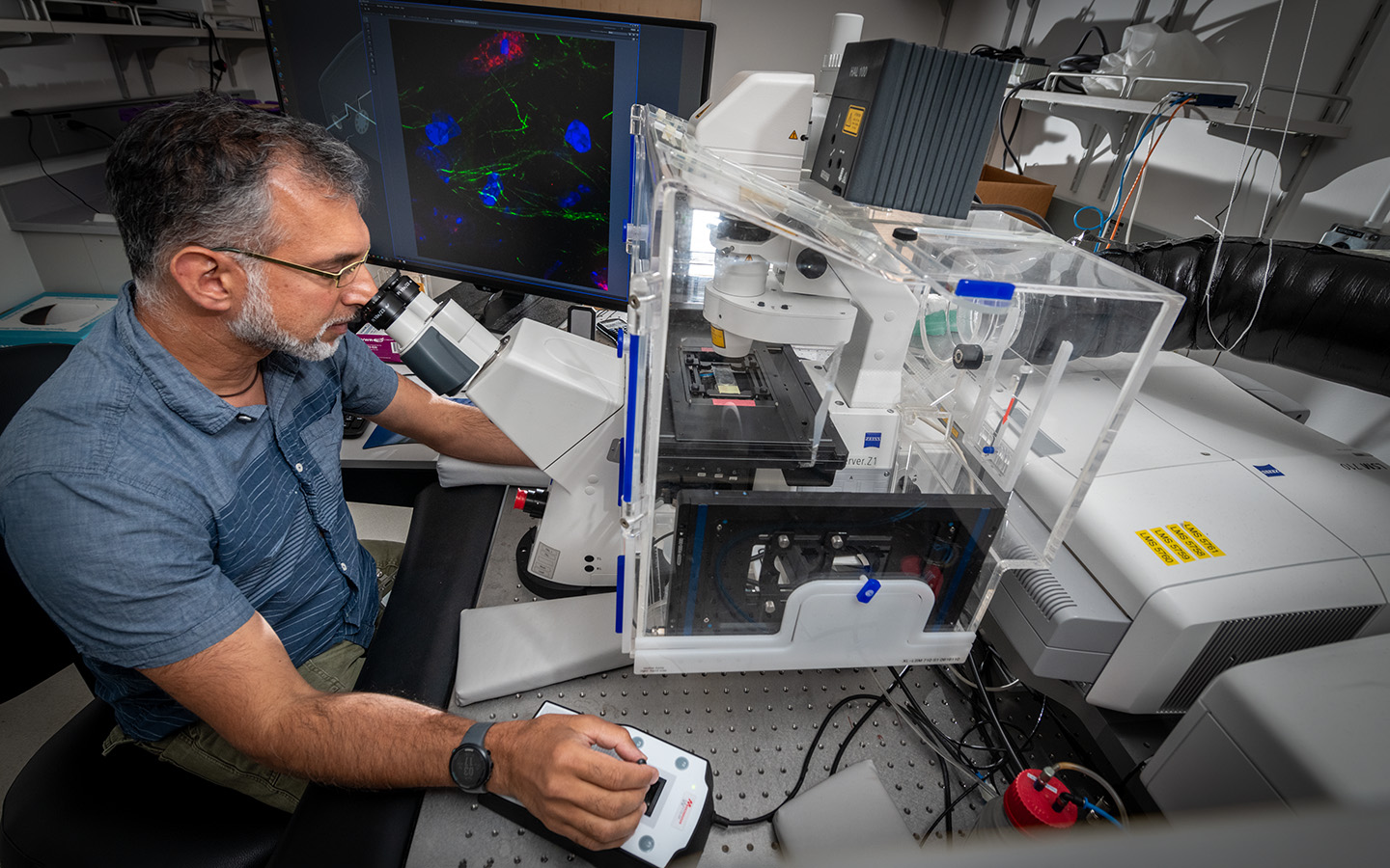 A scientist in a blue shirt looks into a large tabletop microscope. A computer with colorful brain scans is in the background.