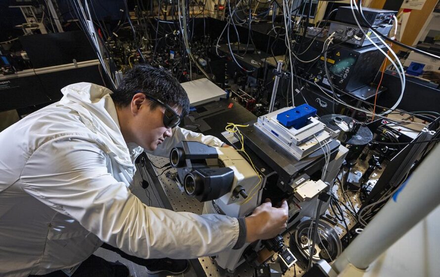 Scientist in protective eyewear adjusts a microscope in a high-tech lab filled with optical and electronic equipment.