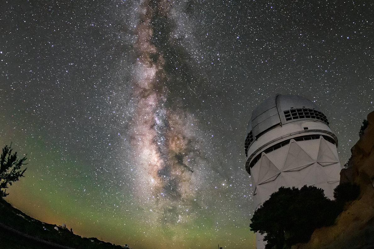 A white structure with a domed top on a rocky outcrop below a star-filled night sky. The Milky Way stretches vertically in a luminous band. Faint green glow tints the curved horizon.