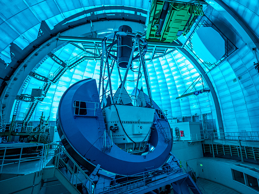 DESI's instrument, a long black cylinder supported by the metal telescope framework, points at the domed ceiling. The dome interior is bathed in blue light.