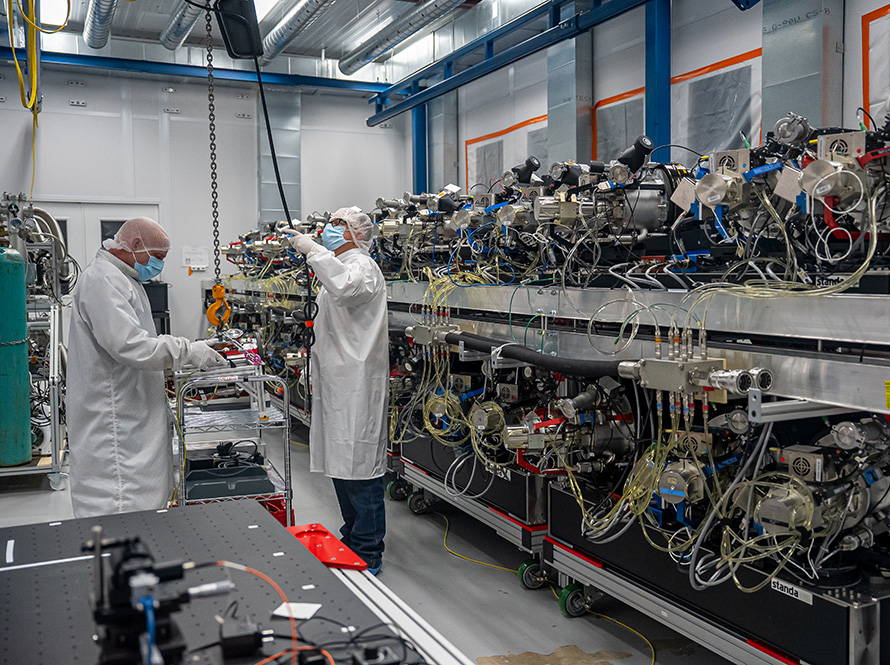 Two people in lab coats, gloves, hair nets, and masks maneuver a hanging hook over a cart. Beside them, equipment with prominent cables is stacked in two rows.