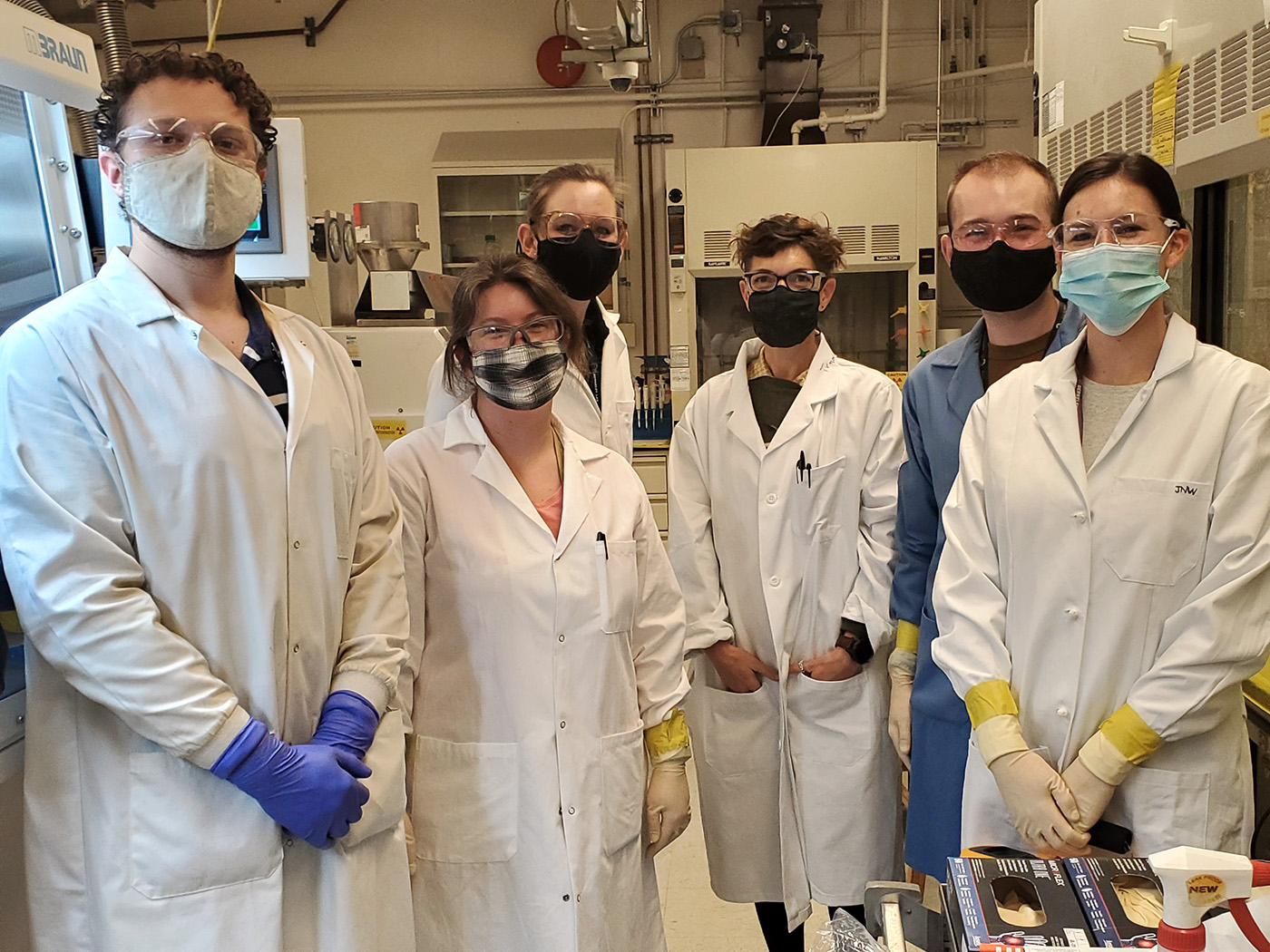 Six researchers in lab coats standing side-by-side in a laboratory.