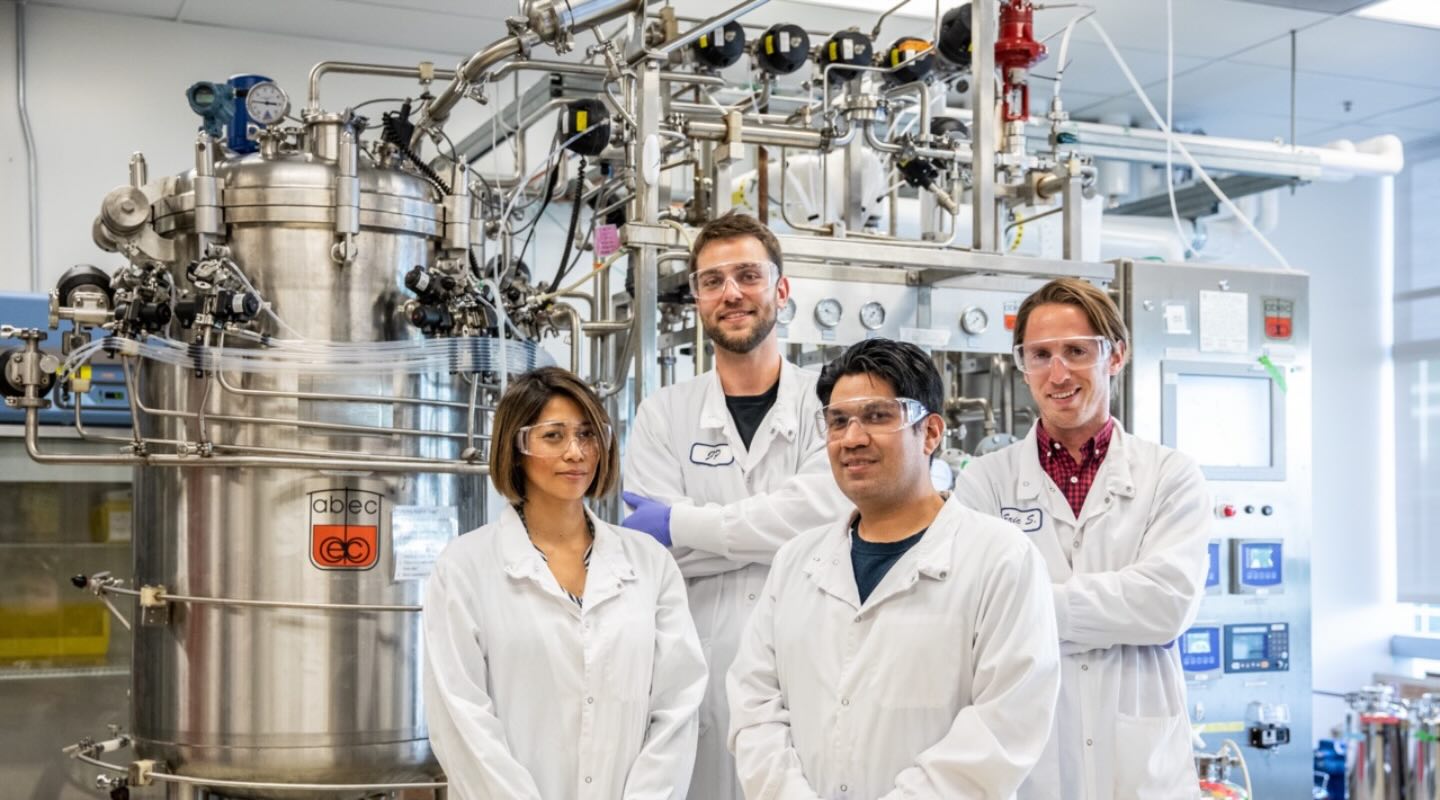 Four scientists with white coats, safety glasses, and blue gloves pose near a large metallic vat attached to a control system with many tubes, valves, and display panels.