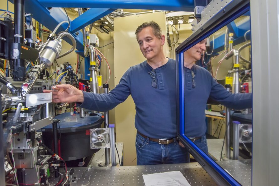 A man in a blue shirt stands smiling next to a metallic instrument with many separate components connected by tubes and wires.