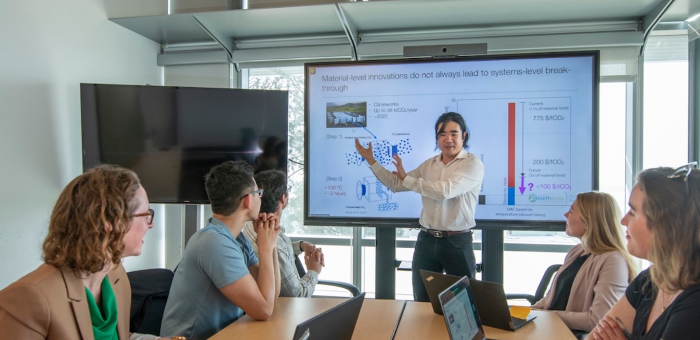 A group of five casually dressed people sit around a table in a conference room, looking at a man presenting at the head of the table, a slideshow with graphics and unreadable text is visible on a large screen behind him.