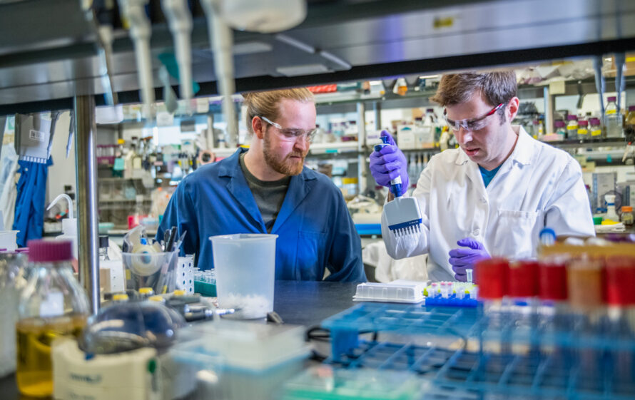 A man in a blue lab coat watches as a colleague in a white lab coat uses a multi-channel pipette in a laboratory. Vials, flasks, and other implements are seen in the foreground, and containers for chemicals fill shelves behind the scientists.